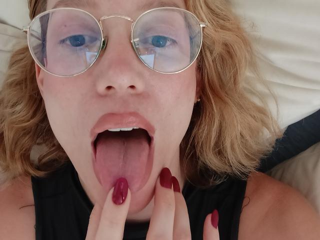 MissFrenchy, avec ses cheveux blonds ondulés, ses lunettes rondes et ses ongles rouges, la langue tirée, touchant ses lèvres avec ses doigts, portant un haut noir, avec des yeux bleus et une langue rose visible..