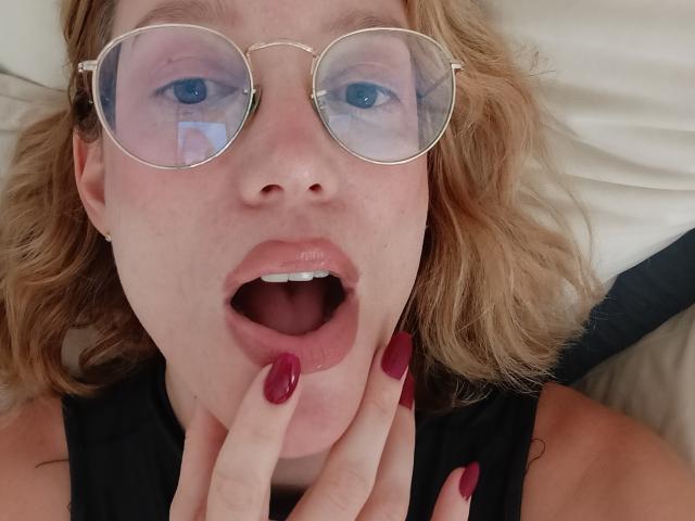 MissFrenchy, usando óculos, com cabelo loiro ondulado e unhas vermelhas, boca aberta, dedos perto do queixo, olhos azuis, blusa preta sem mangas, com uma expressão ligeiramente surpresa..