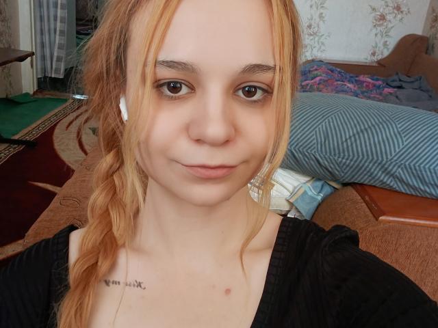 JanelSavala with light blonde hair in a braid, wearing a black top, white earbuds, and a small tattoo near her collarbone, smiling softly at the camera.