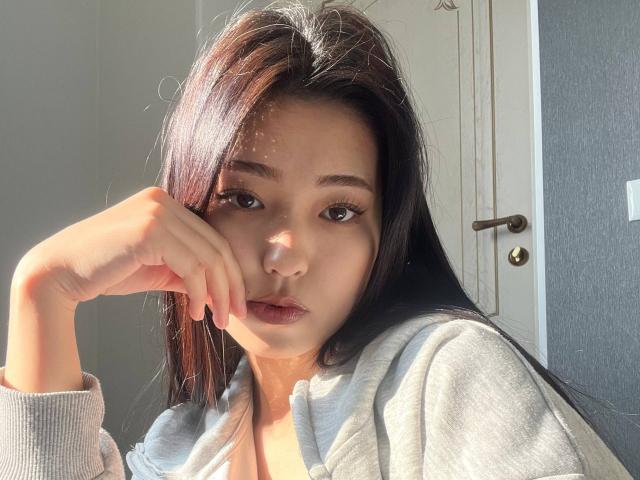 EstherFramer with long brown hair, light skin, wearing a grey hoodie, sunlight on her face, hand resting on her cheek, looking slightly to the side, soft expression.