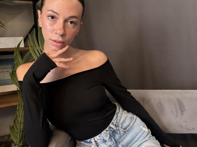 Monnie Karpowich, con le spalle scoperte, indossa una maglietta nera a spalle larghe e jeans blu, con la mano vicino alla bocca, guarda la fotocamera, ha i capelli castani raccolti in uno chignon, la luce è soffusa, la posa è rilassata, il trucco è leggero..