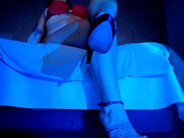 NaniSky lying on a bed, wearing a red bra and white skirt, with legs crossed, showing her tits and black high heels, under blue lighting.