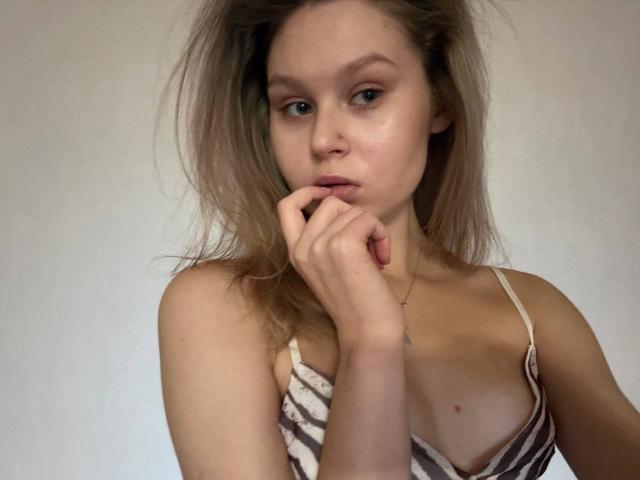 YungQuiller with blonde hair, nose piercing, and a striped top, biting her finger with a serious expression, showing small tits and a slender neck.