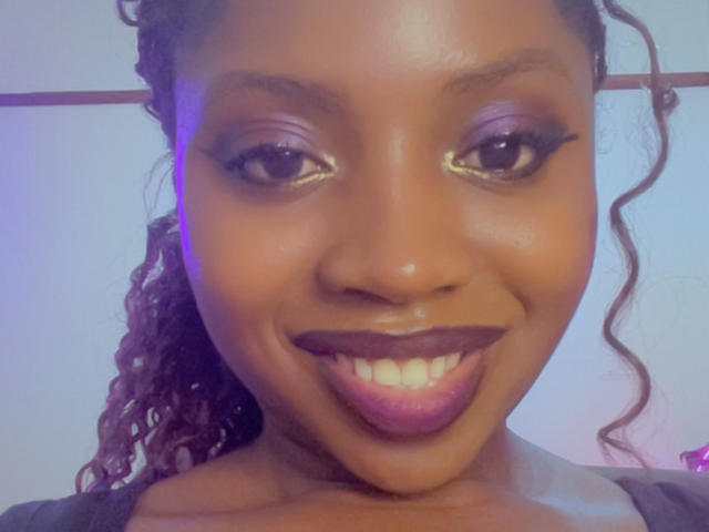 DarkMilanbby with dark brown skin, curly hair, purple eyeshadow, black eyeliner, gold eyeliner, dark lipstick, smiling with visible teeth, wearing a gray top.