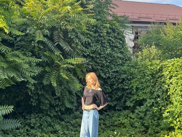 AlineGossom with red hair, black off-shoulder top, and high-waisted blue jeans, standing in lush green ferns, showing her small ass and slightly visible tits.