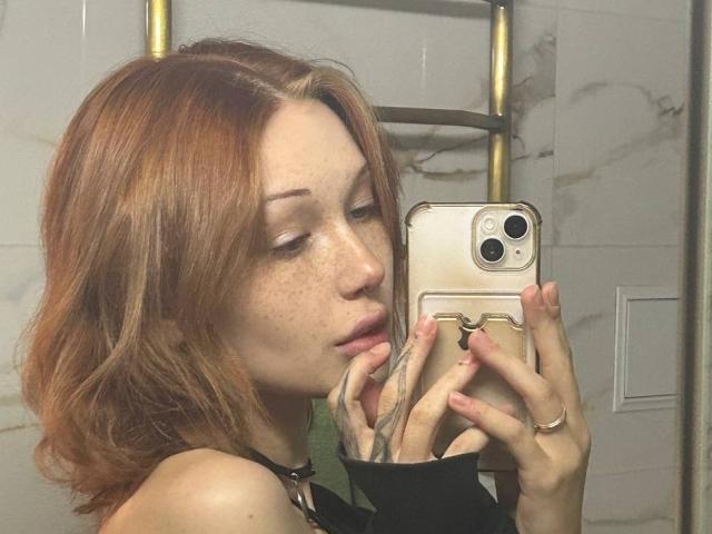 AlineGossom with red hair, freckles, and tattoos, taking a selfie with her phone in a mirror, wearing a black top, showing her nose and lips.