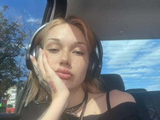 AlineGossom with freckles, pouty lips, nose ring, black headphones, and tattoos, resting her head on her hand in a car, wearing a black top, sunlight on her face.