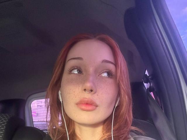 AlineGossom with red hair, nose ring, freckles, and earbuds, pouting in a car, looking sideways, her tits not visible, her ass not visible, in a selfie.