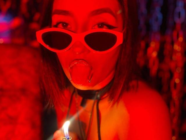 LunaixMoon with red sunglasses, tongue out, and a tongue piercing, wearing a black collar, holding a lighter, in red lighting. Her ass is partially visible.