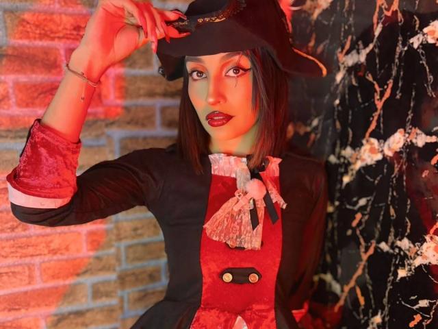 LunaixMoon with dark red lipstick, black hat, and red-black velvet dress, adjusting hat, showing off sharp nails and bracelet, standing against a marble wall.