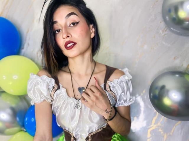 LunaixMoon with dark hair, red lipstick, white off-shoulder top, brown corset, necklace, and hand on chest, surrounded by balloons, has a sexy and cute look.