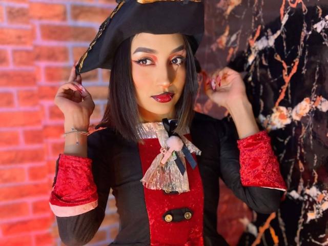 LunaixMoon, a sexy pirate with dark skin, red lipstick, black pirate hat, red velvet cuffs, and a black jacket with a lace bow, touches her hat.