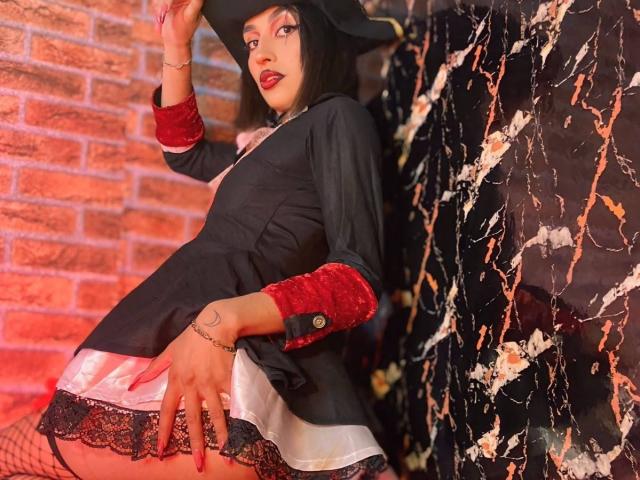 LunaixMoon with dark hair, witch hat, black and red velvet sleeves, black and white dress, fishnet stockings, and lace, touching her ass, red lipstick, brick wall background.