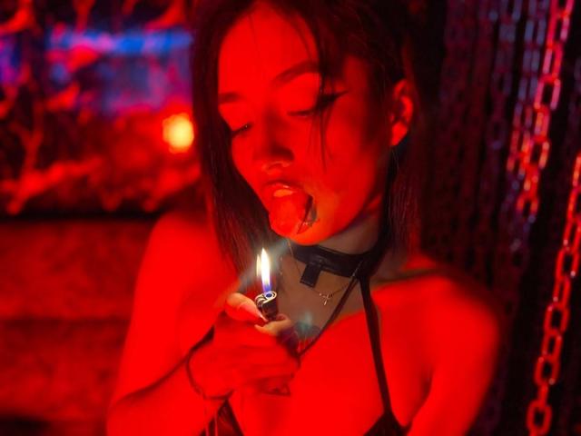 LunaixMoon with red-lit skin, tongue out, holding a lit cigarette, wearing a black choker, and a strapless top, showing her tits, in a dark, chain-filled room.
