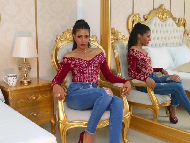 CamilaWest con i capelli scuri raccolti in una coda di cavallo, indossa una maglietta rossa a spalle scoperte, jeans blu, tacchi rossi, seduta su una sedia dorata, il suo riflesso in uno specchio mostra il suo sedere..