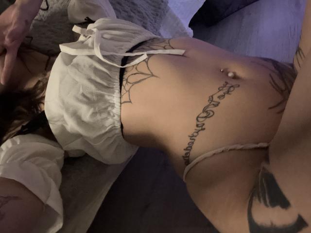 La pancia tesa e tatuata di SimonneBrustkern, con un piercing all'ombelico, indossa una camicia bianca e ampia e uno slip beige, mostrando un tatuaggio di un ragno e un tatuaggio con scritte sotto..