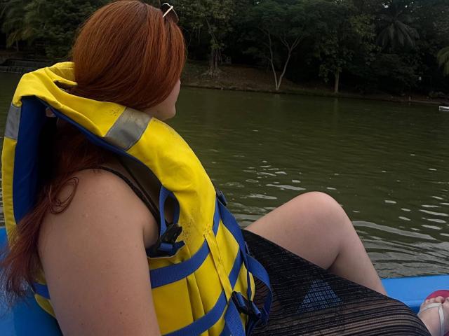 AmberLaurent met rood haar, een gele en blauwe reddingsvest, een zwart gestreepte jurk, zittend op een blauwe boot, met de rug naar de camera gericht, kijkend naar het groene water, met een bebost achtergrond, bewolkt..