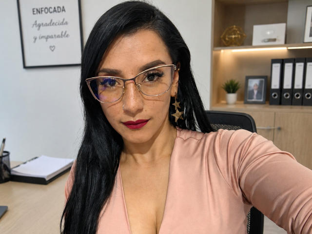 HotCarolinexx met lang zwart haar, een bril, sterrenoorringen en een roze top die de decolleté laat zien, zittend in een kantoor. Rode lippenstift, serieuze uitdrukking, medium huidskleur, aantrekkelijk..