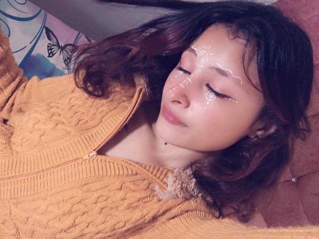 ValentinaRoseX with white face paint, winking, in a yellow knit sweater, lying on a pink cushion. She has dark wavy hair and a small beauty mark on her chin.