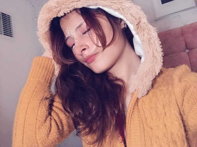 ValentinaRoseX with wavy brown hair, wearing a fluffy beige hood and yellow knit sweater, white face paint, and a pink cushioned headrest in a soft, cozy setting.