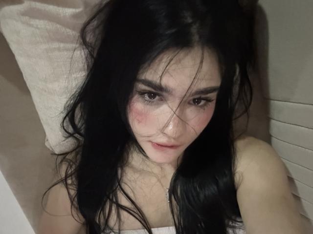 LillyPreaty, con el pelo largo y negro, mirando hacia arriba, tiene mejillas rosadas, pechos pequeños y cejas oscuras, está acostada sobre un cojín, vestida con una blusa blanca, con un aspecto ligeramente despeinado..