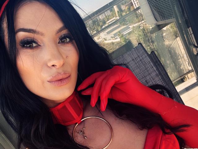 AmelieReea with long black hair, red collar, red gloves, and ring on her tit, looking at the camera, sunlight on her face, city view in the background.