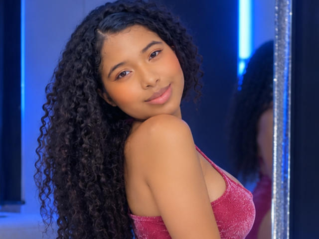 Tifannyy with curly black hair, brown skin, and a slight smile, wearing a pink sparkly top, looking over her shoulder, showing her small, perky tits.