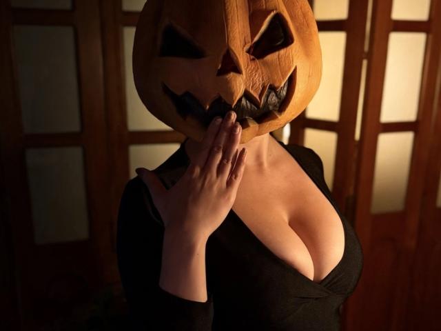 ZoeeMar avec une tête de citrouille sculptée, un haut noir, et une main touchant sa bouche, montrant ses grandes seins nus. Éclairage chaud et tamisé..