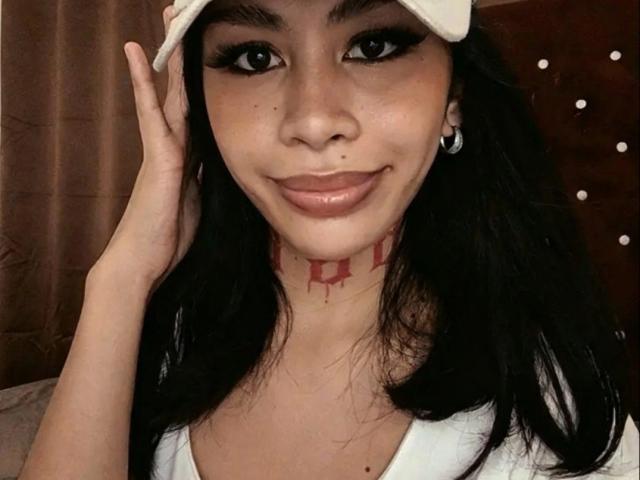MarrahSensual, avec ses cheveux foncés, son bonnet blanc et son maquillage, souriante, tenant sa tête, avec un tatouage rouge sur son cou, un t-shirt blanc et des boucles d'oreilles pendantes, dans un autoportrait..