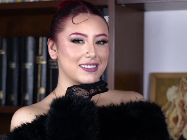 MaddyMills with purple hair, black lace collar, and black furry garment, smiling at camera, showing off her tits, with pink lipstick and eyeliner.