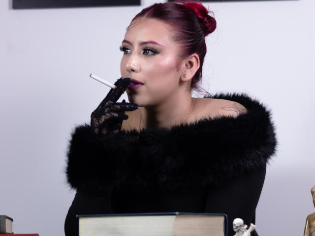 MaddyMills with red hair, black fur-lined top, and lace gloves, smokes a cigarette, looking to the side, showing tattoos on her right shoulder.