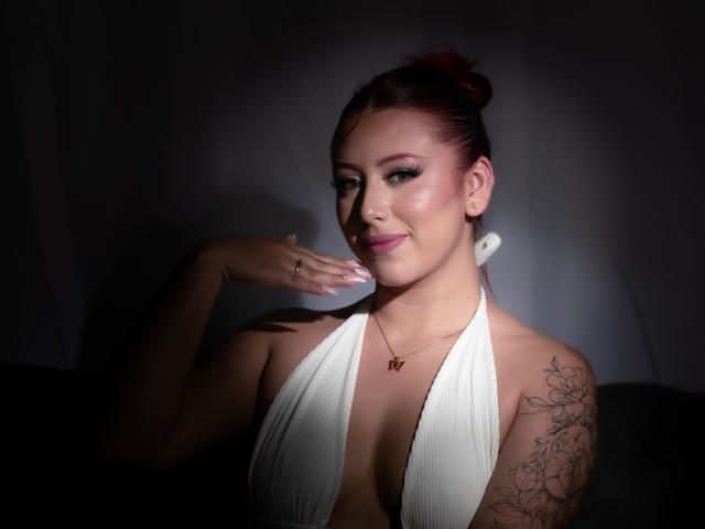 MaddyMills with pink hair in a bun, white halter top, showing tits, tattoo on right arm, hand near face, smiling, necklace, dark background, soft light, confident pose.