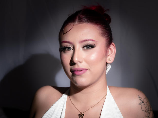MaddyMills has red hair in a bun, white halter top, nose ring, tattoo on left shoulder, pink eyeliner, and a subtle smile. She's wearing a gold necklace.