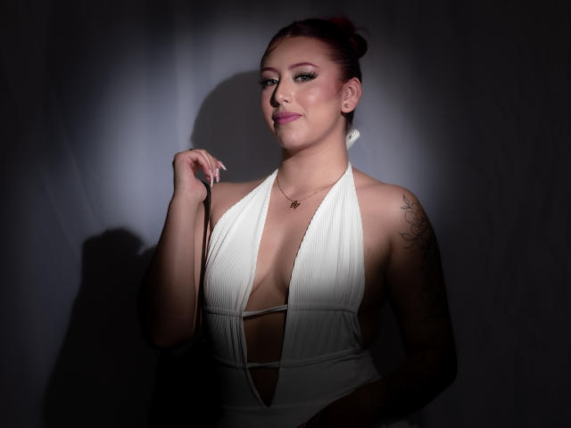 MaddyMills with red hair in a white halter dress, holding a strap, showing cleavage, tattoo on right arm, pink lipstick, shadowed face, and subtle smile.