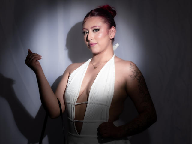 MaddyMills with red hair in a bun, wearing a deep-cut white dress, tattoo on her right arm, holding a black strap, light shadow on her face, smiling.