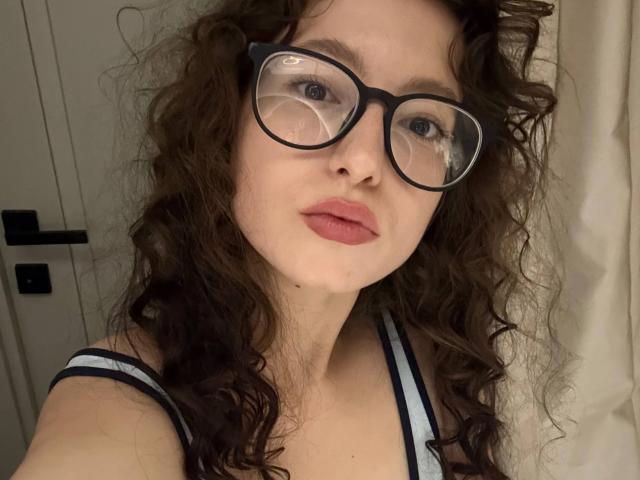 Un selfie de LupeYing con cabello castaño rizado, usando gafas negras y lápiz labial rosa, mostrando su rostro y pecho en una blusa azul a rayas, sosteniendo la cámara ella misma..