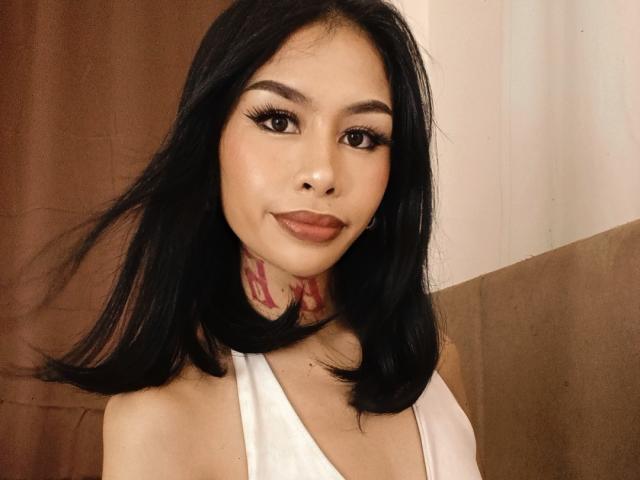 MarrahSensual tiene cabello negro, ojos marrones y un tatuaje en el cuello, lleva una camiseta blanca sin mangas y sonríe con labios carnosos. Sus pechos son ligeramente visibles en la foto..
