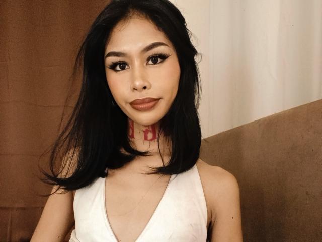 MarrahSensual ha capelli neri di lunghezza media, tratti asiatici e indossa una canotta bianca. Ha un tatuaggio rosso sul collo ed è rivolta verso la fotocamera..