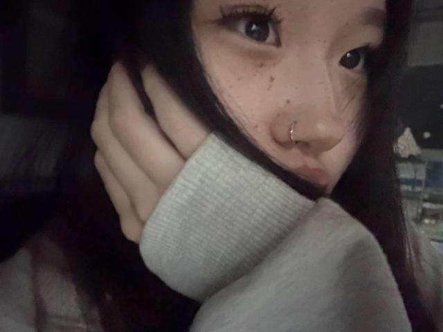 JungMcgranor with dark hair, nose ring, and freckles, holding a grey sweater to her face, showing her black eyes and hand. Her face is partially covered by the sweater.