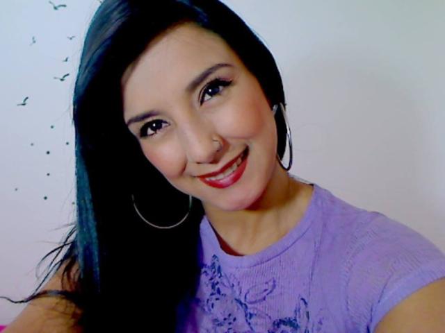 HotCarolinexx with long black hair, smiling, wearing a purple shirt, hoop earrings, and red lipstick, showing her cute nose ring. She's got a sexy, attractive face.