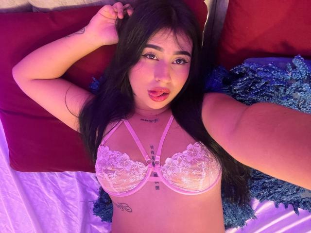 HannahBecker lying on bed, wearing pink lace bra, exposing her tits, with long black hair, pink lipstick, and tattoos on chest, taking selfie with hand above head.