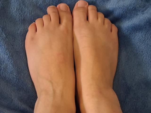 I piedi nudi di Marriam, con le dita leggermente rosate, sono affiancati su una superficie blu e strutturata. La sua pelle è chiara e le unghie dei piedi sono curate..