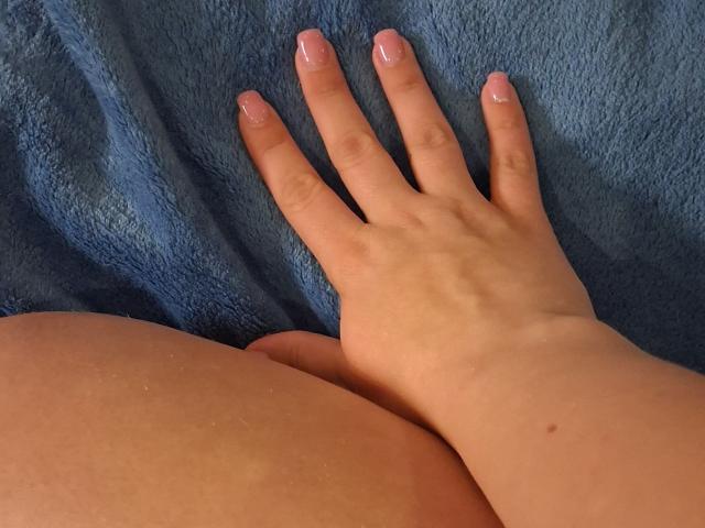 A mão de Marriam, com dedos de unhas rosa, repousa sobre sua nádega nua, e sua pele macia exibe uma textura sutil em contraste com uma toalha azul. O polegar dela aponta para cima, mostrando sua postura relaxada..