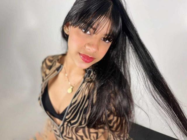 JeanneLacroix with long black hair, brown eyes, and red lipstick, wearing a zebra-patterned top, exposing her small black bra-covered tits. She's smiling slightly and looking up.