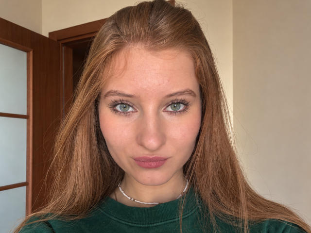 Ardelle Cooksey tiene ojos verdes, cabello rojo largo y labios rosados y gruesos. Lleva un suéter de cuello alto verde y un collar de plata, mirando directamente a la cámara..