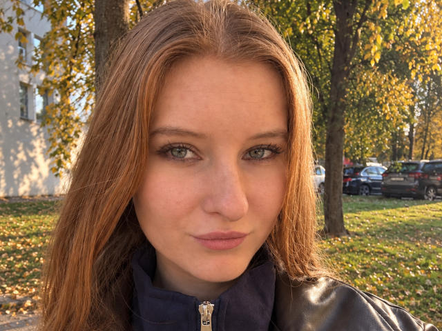 ArdelleCooksey has light green eyes, long brown hair, and a slight pout, wearing a black leather jacket with a zipper. She's outdoors, looking directly at the camera.