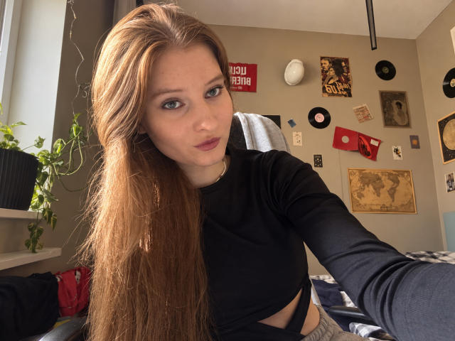 Ardelle Cooksey, met lang, recht bruin haar, draagt een zwart topje met lange mouwen dat haar borsten laat zien, en maakt een selfie in een kamer met decoratie aan de muur en een plant..