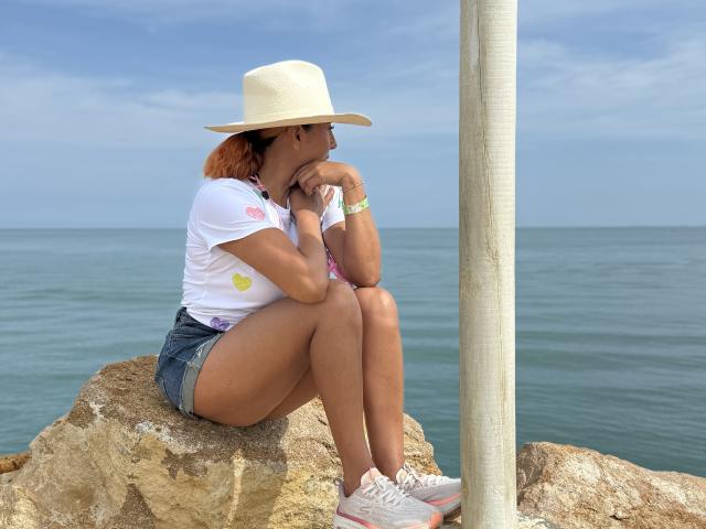 NikiiFoxy, sitting on rocks by the sea, wearing a white hat, white tee, denim shorts, and pink sneakers, with her chin on her hand, looking off to the side.