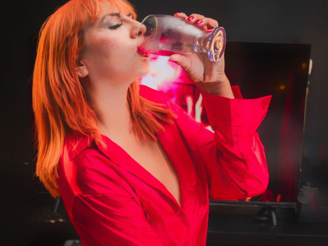 NikiiFoxy con cabello naranja, camisa roja, bebiendo de una botella transparente, líquido rosa salpicando, ojos cerrados, sosteniendo la botella con la mano derecha, esmalte de uñas rosa, busto mediano..