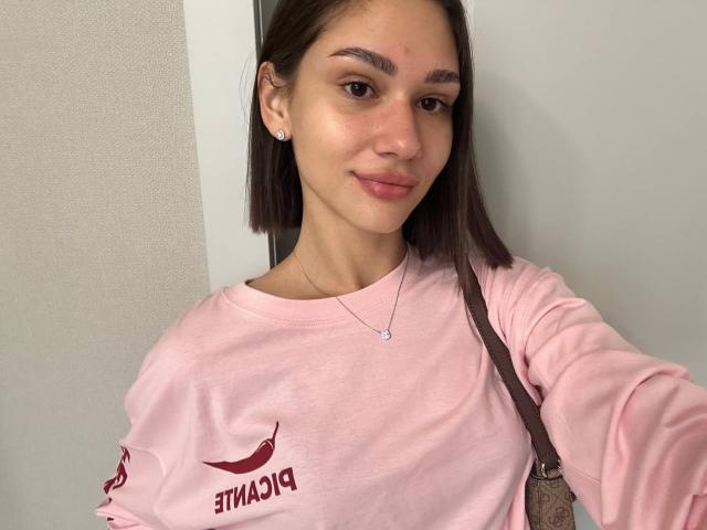 TobiQuinby a un joli visage avec des cheveux bruns lisses, elle porte une chemise rose et un collier, et tient un sac marron, avec une petite imperfection sur le front..